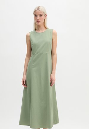 Femme blonde portant une robe sans manches, longue jusqu'aux chevilles, de couleur vert clair, debout devant un fond blanc uni.