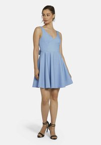Helles blaues ärmelloses Kleid mit V-Ausschnitt, tailliertem Oberteil und ausgestellt Rock. Hergestellt aus strukturiertem Stoff, kombiniert mit schwarzen Riemchenheels.