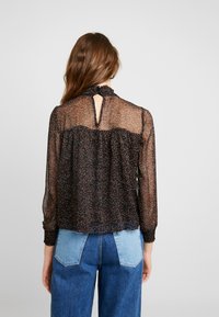 Blusa nera trasparente con colletto a volant, caratterizzata da una schiena con apertura a chiave, maniche lunghe e macchie multicolori diffuse. Indossata sopra dei jeans.