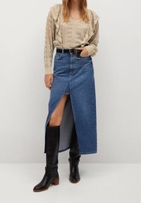 Kobieta ubrana w beżowy sweter z dzianiny, niebieską spódnicę dżinsową z wysokim stanem i rozporkiem z przodu, czarny pasek i czarne wysokie botki do kolan, stojąca na jednolitym tle.