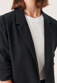 Blazer nero con un design strutturato, caratterizzato da un colletto a un bottone, vestibilità su misura e texture morbida, indossato sopra una maglietta bianca.