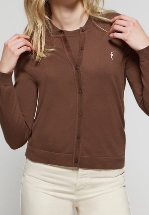 Femme portant un cardigan marron boutonné avec un petit logo blanc brodé sur la poitrine et un pantalon couleur crème.