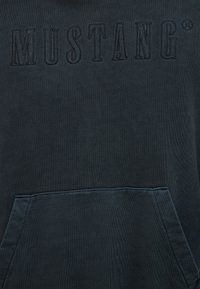 Tmavomodrá mikina s textúrovaným povrchom, veľkým nápisom "MUSTANG" vyšitém v hornej časti a predným vreckom v páse.