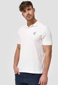 Weißes Poloshirt aus strukturiertem Baumwollstoff, mit Kragen, kurzen Ärmeln und einem kleinen gestickten Logo auf der linken Brust.