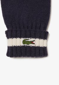 Puño de suéter de punto en navy con textura de canalé y rayas horizontales en crema, que presenta un logo de cocodrilo verde con acentos rojos.