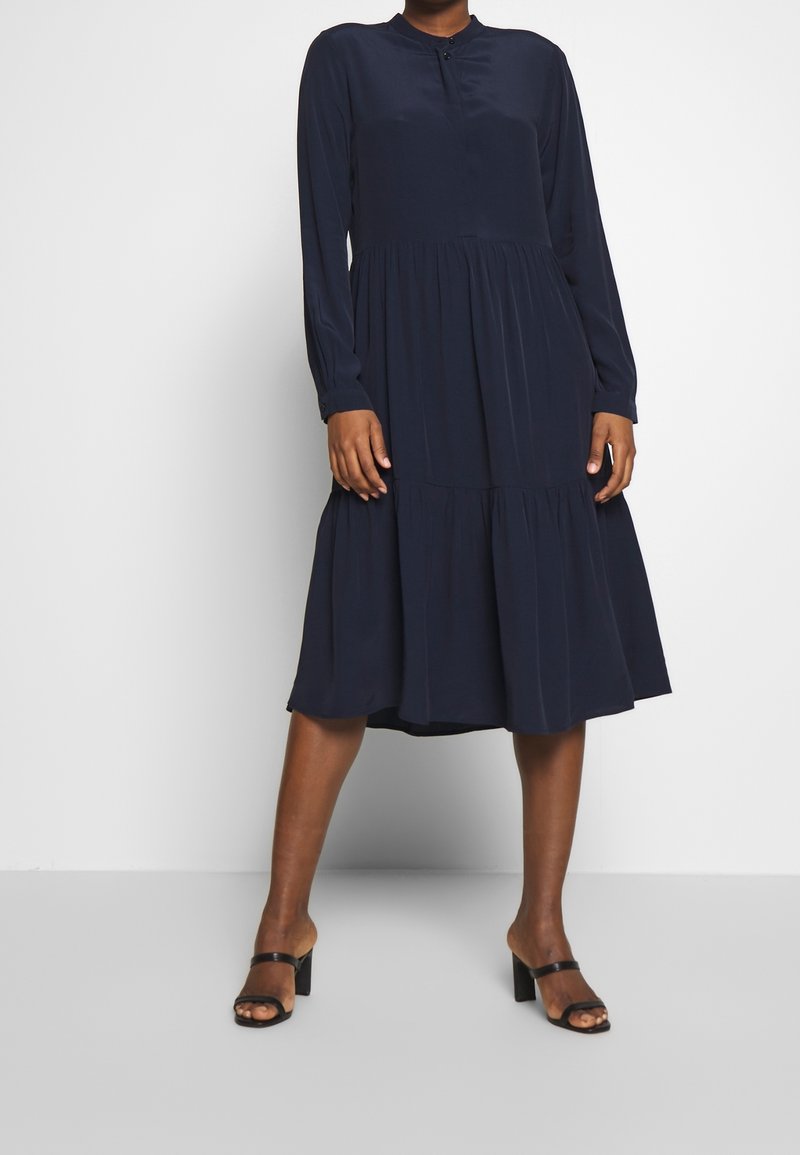 Personne portant une robe midi à manches longues, bleu marine, à étages, associée à des sandales noires à talons, debout devant un fond gris clair uni.