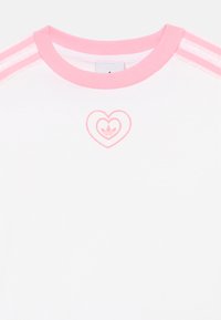 Fehér póló rózsaszín szegéllyel és három rózsaszín csíkkal minden vállon. A mellkason rózsaszín szív alakú logó látható.