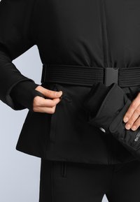 Crna izolirana jakna s teksturiranim strukom, s patentnim džepom, i ruka koja drži presavijenu crnu rukavicu blizu šava.