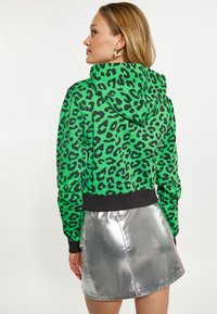 Hoodie cropped con stampa leopardata verde, con polsini e orlo neri, abbinato a una mini gonna luminosa argento. Tessuto testurizzato con una finitura liscia.