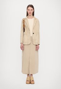 Beige puuvillablazer, jossa on yksi nappi, paritettuna samasta kankaasta valmistetun pitkän hameen kanssa. Beige platform-sandaalit, luonnollisen värinen olkalaukku hihnalla.