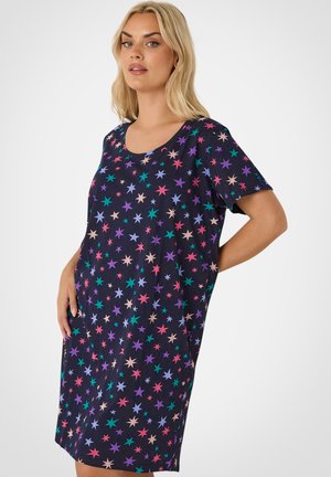 Kobieta ubrana w luźną, granatową sukienkę z krótkim rękawem i kolorowym wzorem gwiazd, stojąca z jedną ręką na biodrze.