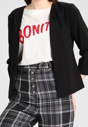 Blazer noir à manches trois-quarts, associé à un t-shirt blanc avec du texte rouge, et un pantalon à carreaux taille haute avec des détails de boutons.