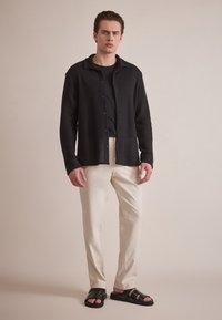 Svart, teksturert skjorte med knapp foran over en svart t-skjorte, kombinert med lyse beige bukser og svarte sandaler. Enkel design og nøytrale farger.