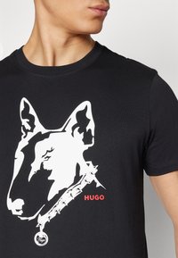 T-shirt noir avec un graphique de bullterrier blanc et le texte "Hugo" en rouge. Tissu doux, col rond, manches courtes et détail distinct au col.