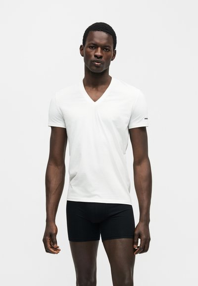 Άντρας που στέκεται φορώντας λευκό μπλουζάκι με V-λαιμόκοψη και κοντά μαύρα εφαρμοστά σορτς μπροστά σε λευκό μονόχρωμο φόντο.