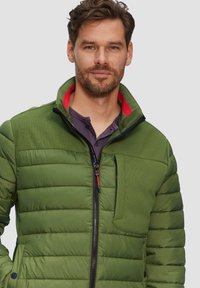 Grüne gepolsterte Jacke mit hohem Kragen, ausgestattet mit einem Reißverschluss vorne und einem strukturierten Schulterbereich. Rote Akzente in der Nähe des Kragens.