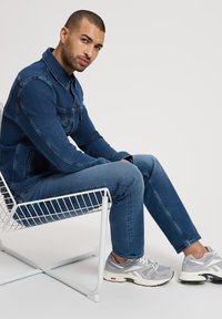 Jeansowa kurtka i dżinsy w średnim niebieskim odcieniu, o dopasowanym kroju. Noszone z szarymi butami sportowymi, siedząc na białym drucianym krześle.