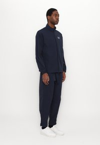 Giacca leggera blu navale con zip frontale, colletto alto e pantaloni abbinati. Le sneakers bianche completano l'outfit, sfoggiano un elastico in vita.