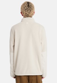 Lys beige fleecejakke med lange ærmer, høj krave og et logo mærke på ryggen. Glat tekstur, afslappet pasform.