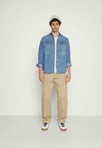 Νεαρός άνδρας με μπεζ καπέλο, μπλε τζιν πουκάμισο πάνω από λευκό μπλουζάκι, μπεζ παντελόνι και λευκά αθλητικά παπούτσια, στέκεται μπροστά σε ανοιχτό χρώμα φόντο.