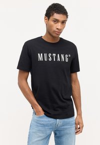 Melna kokvilnas T-krekls ar apaļu kakla izgriezumu un īsām piedurknēm, uz krūšu daļas ir balts teksta uzraksts "MUSTANG". Saskaņots ar gaiši ziliem džinsiem.