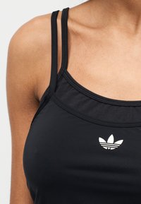 Μαύρο αθλητικό τοπ με λεπτές τιράντες, στρογγυλή λαιμόκοψη και λευκό λογότυπο Adidas στο στήθος. Λείο ύφασμα με εφαρμοστή σιλουέτα.