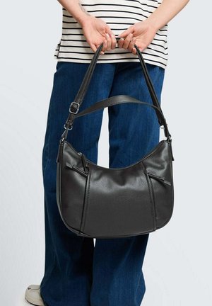 Persona che tiene una borsa nera in pelle con tracolla regolabile dietro la schiena, indossando una camicia a righe e jeans blu.