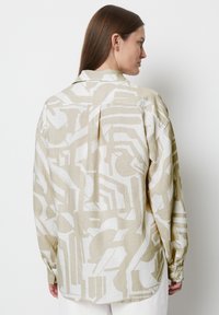 Beige linen shirt with abstract white geometric patterns, relaxed fit, button cuffs, and back pleat detail, shown from behind.