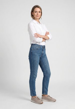 Žena stojící se založenýma rukama, v bílém tričku, modrých džínách a béžových teniskách na jednobarevném světlém pozadí.