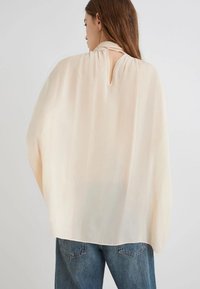 Blusa leggera beige chiara con schiena a chiave, design drappeggiato e maniche larghe. Realizzata in un tessuto morbido, abbinata a jeans in denim blu.