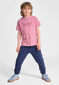 Różowy bawełniany t-shirt z wytłoczonym logo, zestawiony z granatowymi dresami i jasnoniebieskimi sneakersami z białymi akcentami. Luźny fason.