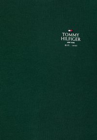 Camiseta de algodón verde oscuro con texto blanco que presenta el logo de "TOMMY HILFIGER", junto con "NEW YORK" y "EST. 1985" debajo.
