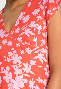 Robe corail avec un décolleté en V profond, ornée de motifs floraux roses et de manches courtes. Le tissu semble léger avec une texture lisse.