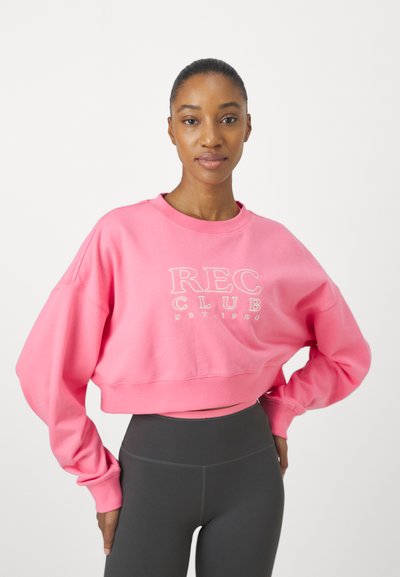 Rózsaszín cropped pulóver széles ujjal, fehér "REC CLUB" logóval. Sötétszürke magas derekú leggings-szel párosítva.