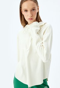 Sudadera de color crema claro con diseño de media cremallera, capucha ajustable con cordón y tejido suave y liso; presenta hombros caídos y un ajuste relajado.