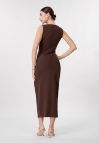 Γυναίκα με τα μαλλιά σε κότσο φοράει μίντι καφέ φόρεμα χωρίς μανίκια και μπεζ πέδιλα με ψηλό τακούνι, πόζαρε με το ένα χέρι στη μέση μπροστά σε λευκό φόντο.