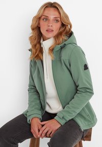 Green softshell jacket with a hood, zip pockets, and black logo patch. Worn over a white fleece top, paired with dark jeans.