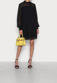 Robe noire à manches longues avec un motif en zigzag, accompagnée d'un sac à main jaune au design structuré et aux ferrures dorées. Talons noirs à brides.