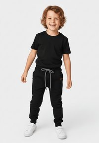 Uśmiechnięte dziecko o kręconych włosach stojące, ubrane w czarny t-shirt, czarne spodnie dresowe ze sznurkiem oraz białe trampki na jednolitym tle.