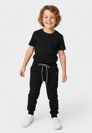 Uśmiechnięte dziecko o kręconych włosach stojące, ubrane w czarny t-shirt, czarne spodnie dresowe ze sznurkiem oraz białe trampki na jednolitym tle.