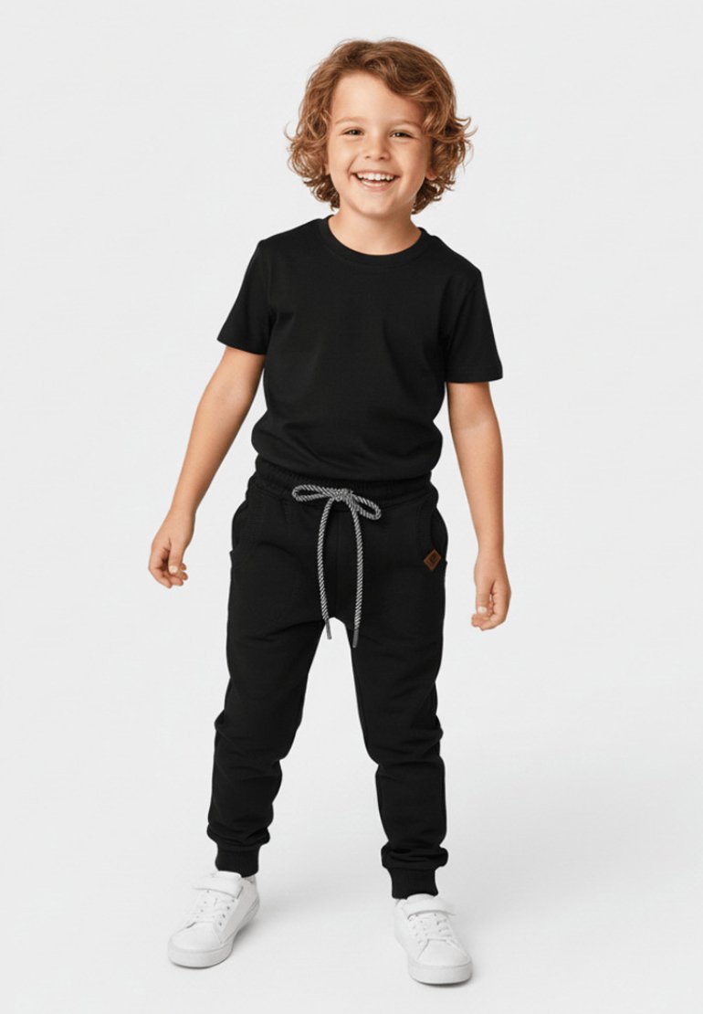 Uśmiechnięte dziecko o kręconych włosach stojące, ubrane w czarny t-shirt, czarne spodnie dresowe ze sznurkiem oraz białe trampki na jednolitym tle.