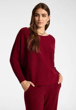 Vrouw draagt een loszittende bordeauxrode trui en broek, met lang bruin haar, gouden ketting, oorbellen en armband, handen in de zakken.