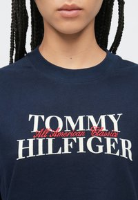 Μπλε σκούρο βαμβακερό t-shirt με το λεκτικό "TOMMY" σε λευκό και "HILFIGER" σε λευκό, με την επιγραφή "All American Classics" σε κόκκινη καλλιγραφία από κάτω.