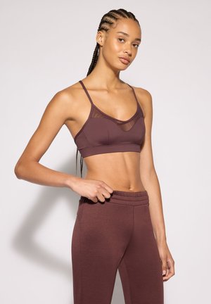 Donna con capelli intrecciati che indossa un reggiseno sportivo marrone e leggings abbinati a vita alta, posando su uno sfondo chiaro uniforme.