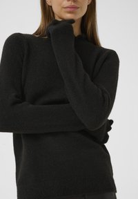 Žena oblečená v černém svetr s dlouhým rukávem a texturovaným materiálem, pózující s jednou rukou přes trup a druhou rukou dotýkající se brady.