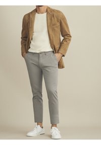 Mies, který má na sobě béžovou suedenou blazeru, bílý texturovaný svetr, šedé slim-fit kalhoty a bílé tenisky, s rukama v kapsách.