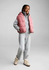 Vesta roz căptușită peste un pulover tricotat gri, blugi gri deschis și cizme bej cu bordură din blană. Modelul stă cu o postură încrezătoare.