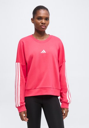 Femme portant un sweat-shirt rose à manches longues avec des bandes blanches sur les manches et des leggings noirs, debout devant un fond clair uni.