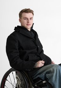 Schwarzer Wollmantel mit Reverskragen und Knopfverschluss, getragen über einer schwarzen, mit Reißverschluss versehenen Innenjacke, kombiniert mit blauen Jeans. Rollstuhl im Bild.