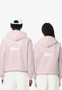 Sudaderas con capucha rosas que presentan el logo en blanco de "LACOSTE" y el cocodrilo en la parte posterior. Pantalones blancos de tejido texturizado y diseño de corte relajado.